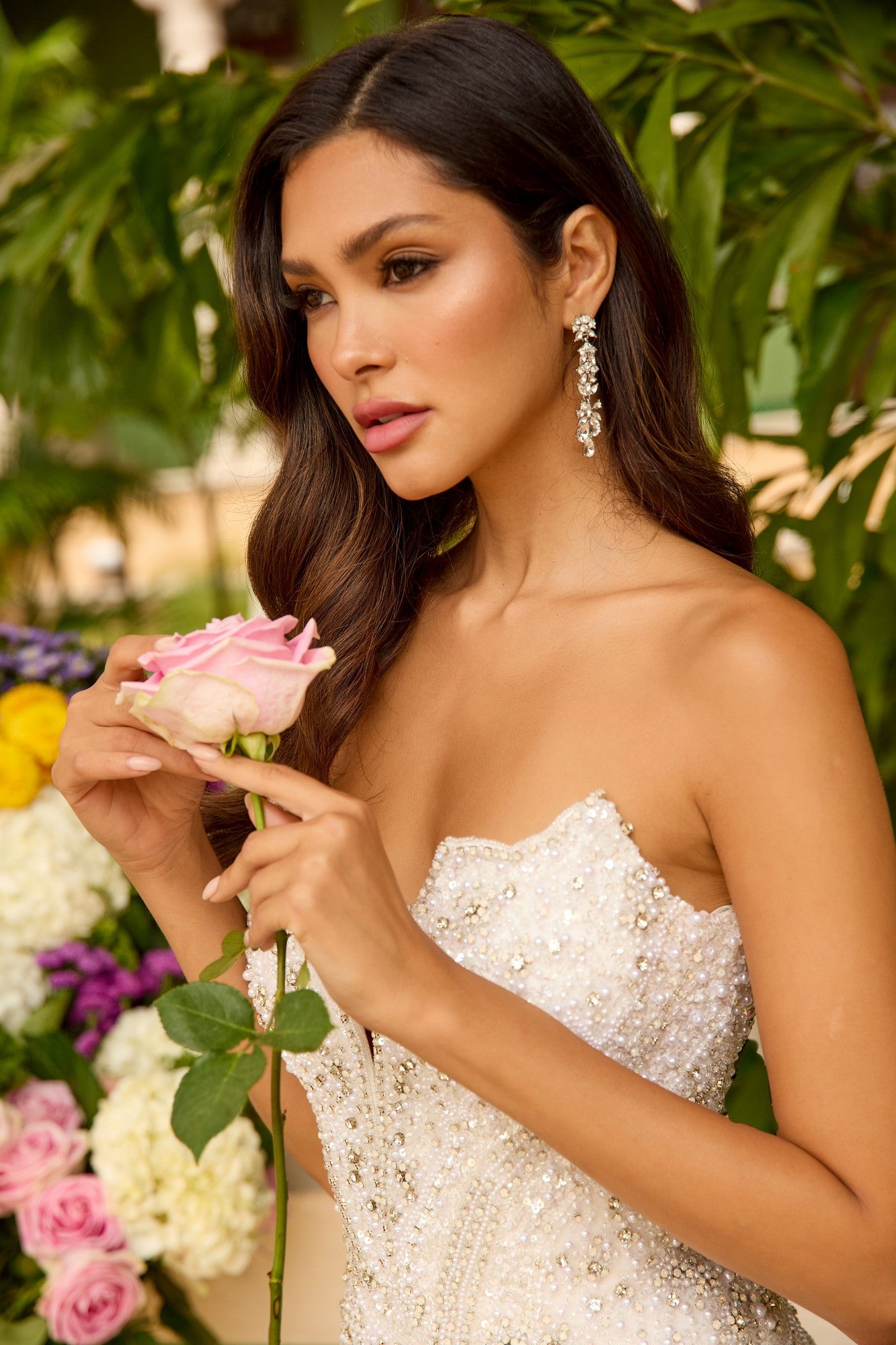 A woman in the Ava Presley 47244 long prom dress holds a pink rose, standing before green leaves and colorful flowers, gazing thoughtfully to the side.